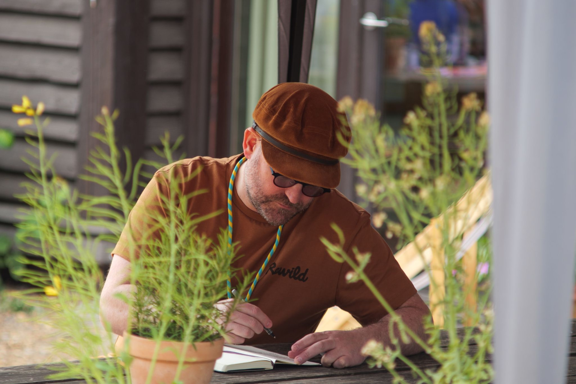 A man in a brown cap writing in a notebook at an outdoor wooden table